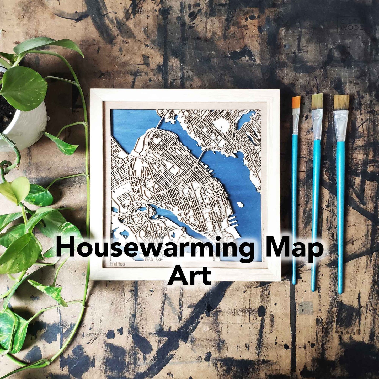 Framed laser-cut wooden map of Halifax, with teal-handled paintbrushes and a potted plant on a splattered tabletop.
